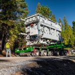 A specialized transportation crew member operates an SPMT carrying a transformer.