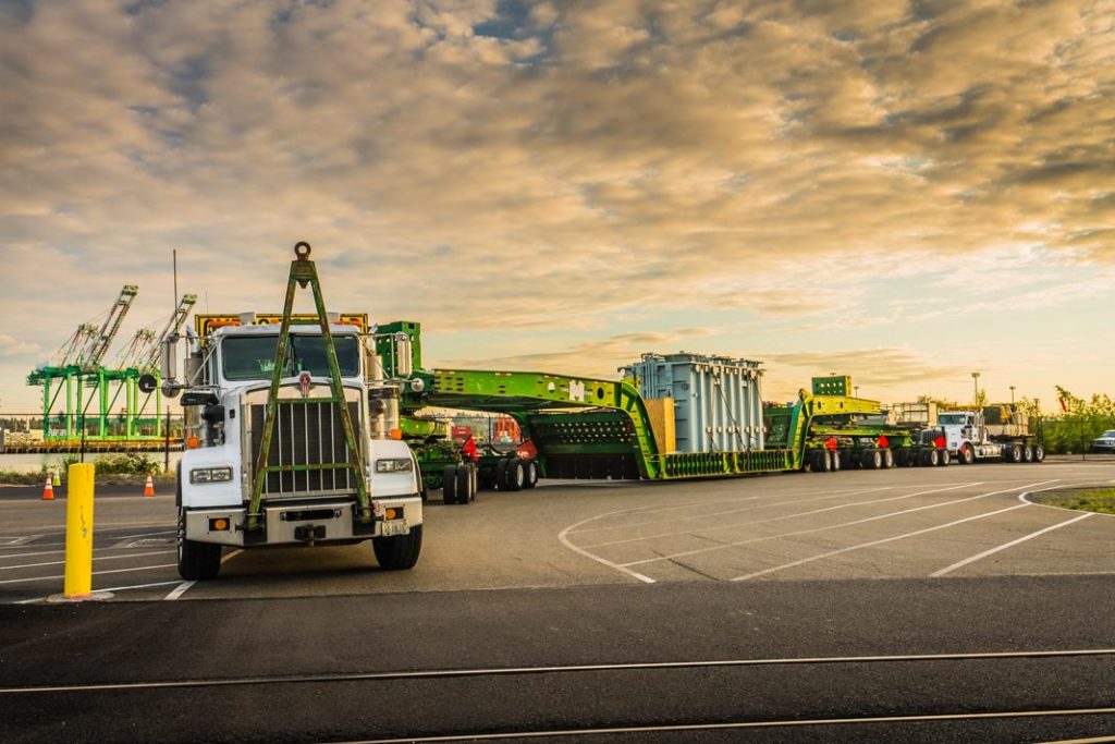 2015 Kenworth C500 truck parked on pavement with a 150-ton trailer carrying a shunt reactor in front of a cloudy sky